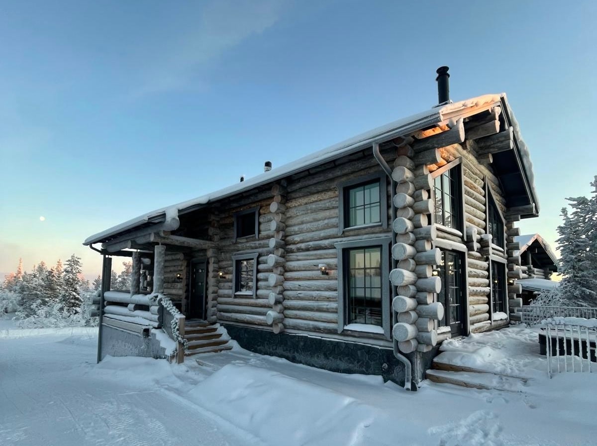The exterior of a log cabin is presented, characterized by its rustic architecture and snow-covered structure. Tall windows are framed by natural wood, allowing light to filter in. Snow blankets the surrounding landscape, enhancing the cabin's charm in a serene winter setting.