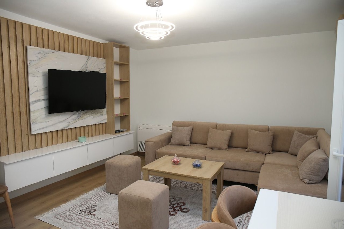 A contemporary living room is presented, featuring a large, L-shaped sofa in neutral tones. A wooden coffee table sits in front, accompanied by smaller cushioned stools. A flat-screen television is mounted on a light-colored wall, complemented by wooden shelving and soft overhead lighting.