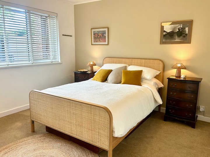 Elegant bedroom with rattan bed frame, white bedding, and yellow accent pillows. Dark wood nightstands with table lamps, natural light from window blinds, and warm neutral tones create comfortable, inviting space. Classic bedroom design.
