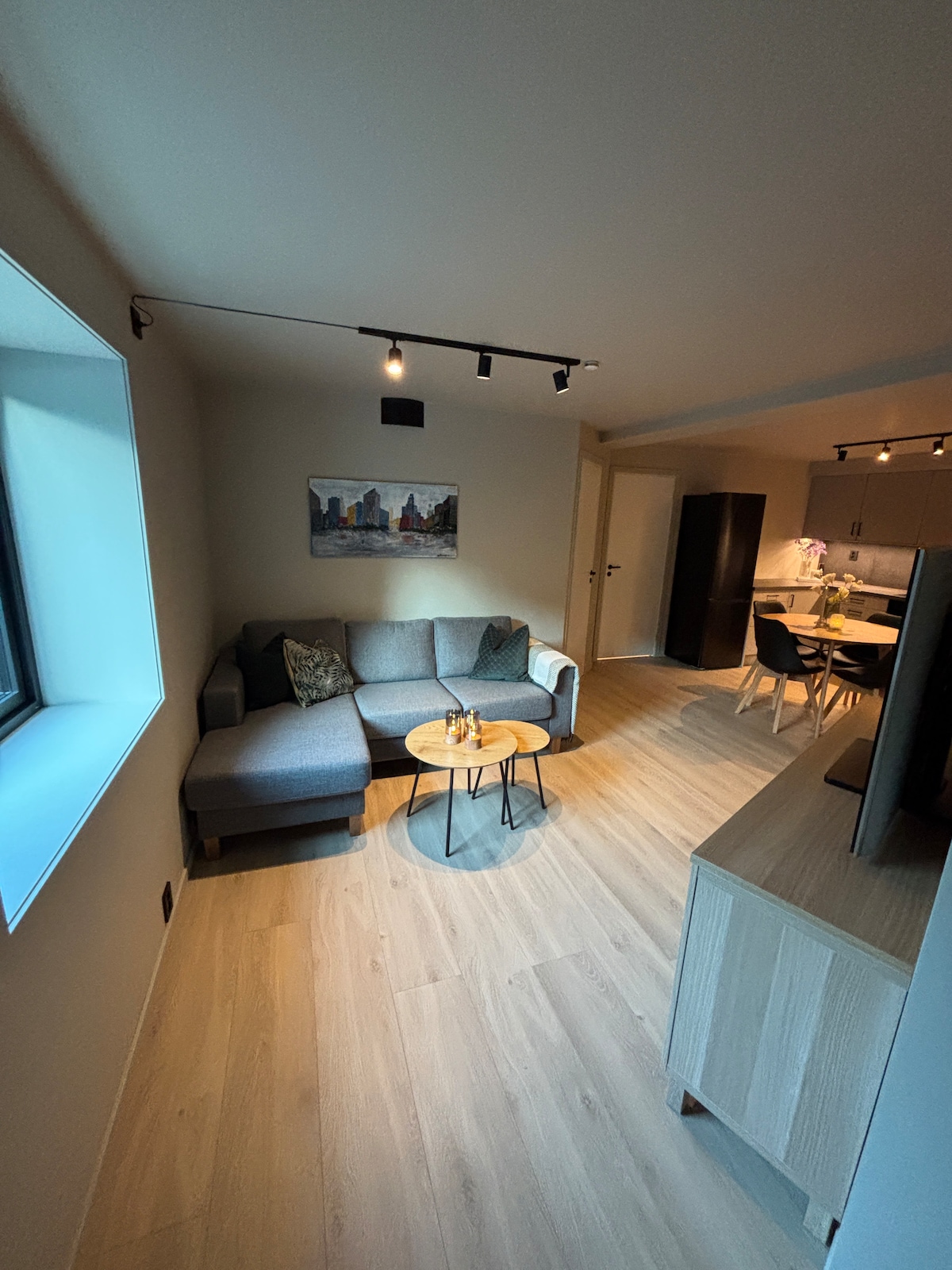 A comfortable seating area is featured, consisting of a grey sectional sofa with decorative pillows. A small round table sits in front of the sofa. A painting depicting cityscapes is displayed on the wall, with soft, natural light coming through a large window.