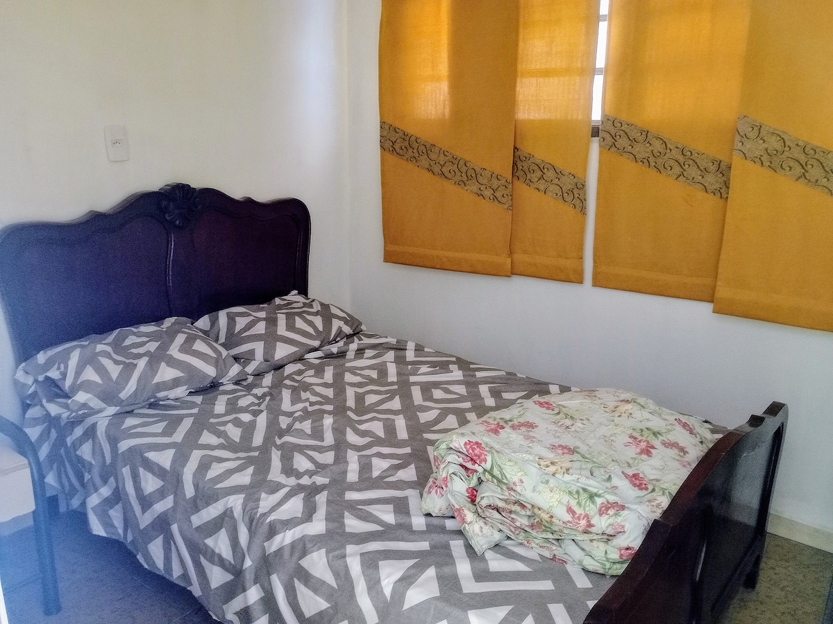 A compact sleeping area features a double bed with a geometric-patterned duvet and floral blanket. The walls are adorned with light-colored curtains, partially covering a window. A simple chair is placed beside the bed, contributing to the overall functionality of the space.
