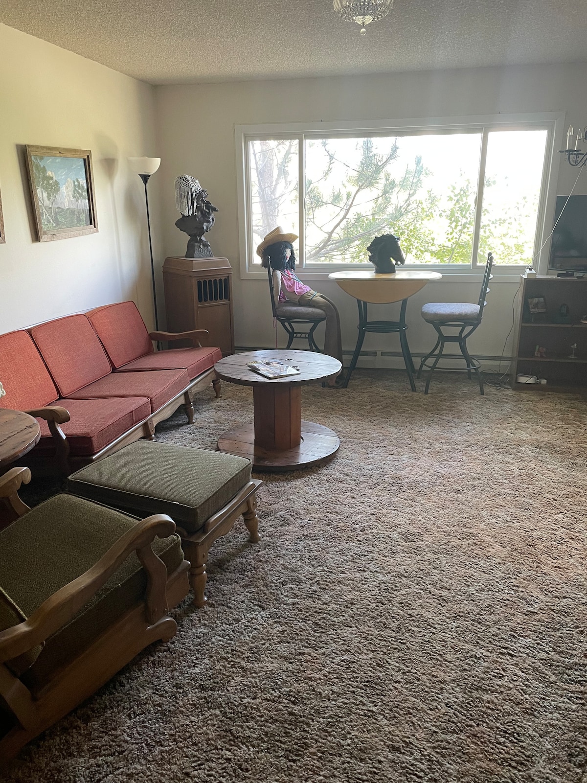 A cozy living space features a light brown carpet and an array of seating options, including a red sofa and two armchairs. A round wooden table is positioned near a large window, allowing natural light to illuminate the room. Decorative paintings adorn the walls.