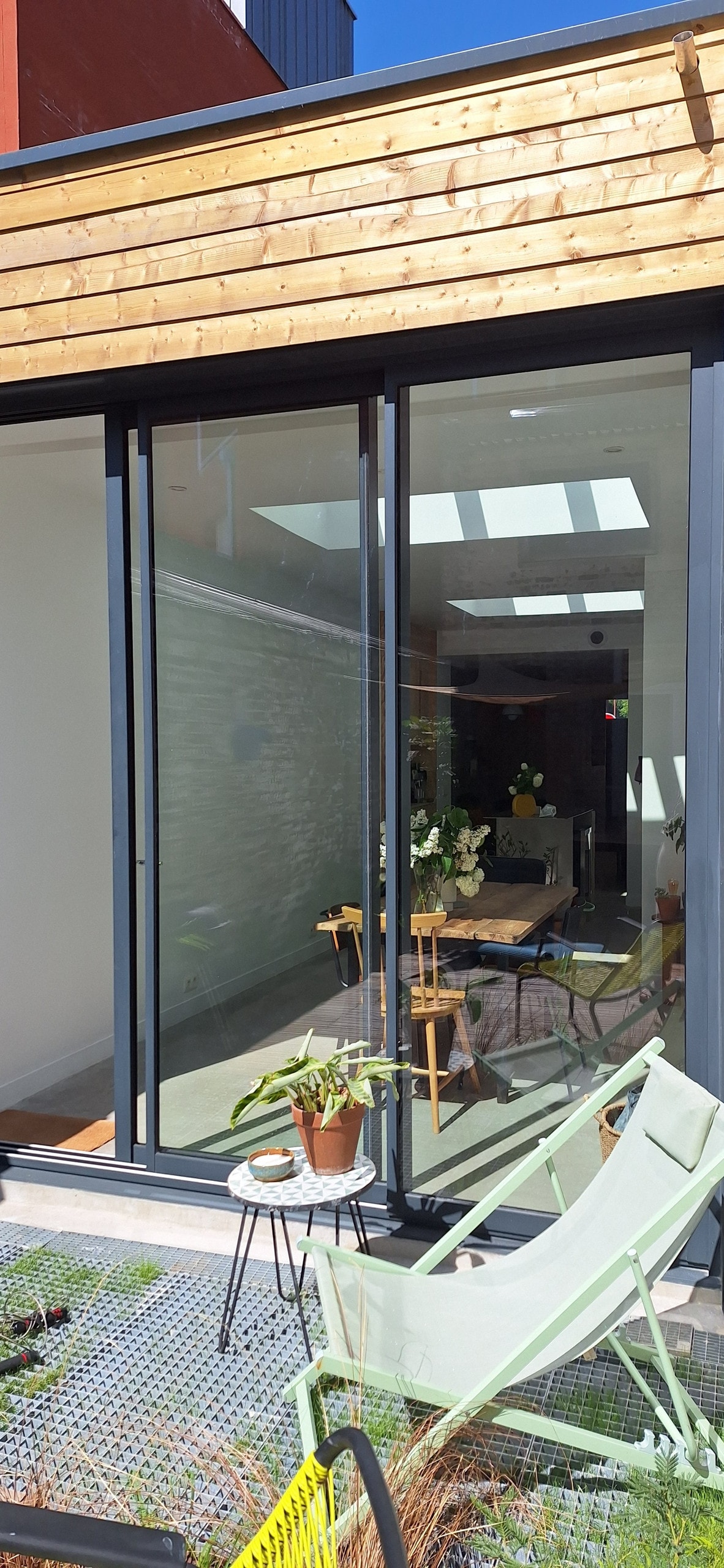 A bright and airy interior space is visible through expansive glass sliding doors. A dining table with chairs is set against a plant-adorned area. Natural light filters in through a skylight, enhancing the open layout and connection to the outdoors.