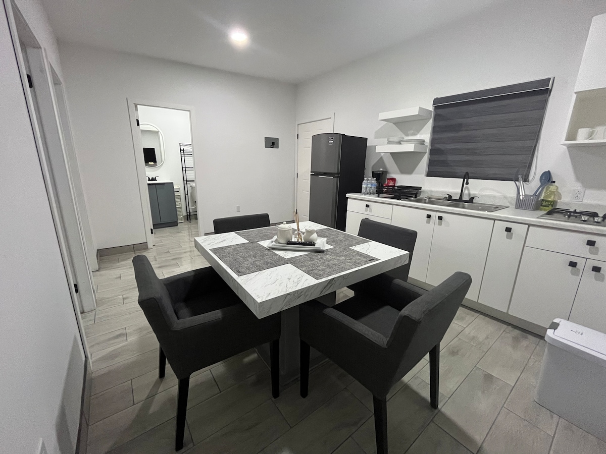 A modern kitchen area is presented with a marble dining table surrounded by four upholstered chairs. A refrigerator and kitchen cabinets are visible, alongside essentials on the countertop. Soft lighting complements the bright, neutral walls and tiled flooring.