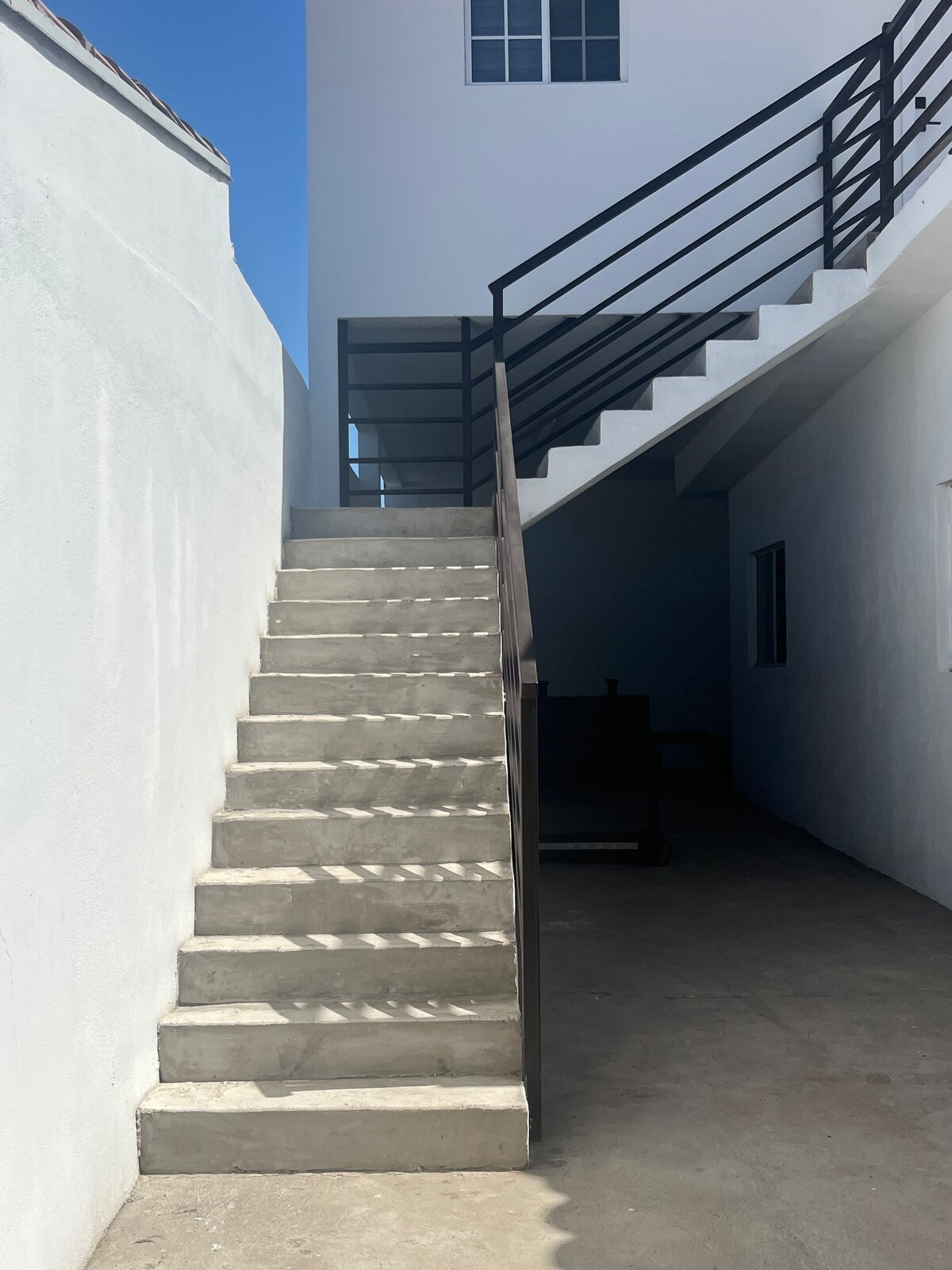 A clean, concrete staircase leads upwards, flanked by white walls and a black railing. Natural light illuminates the steps, casting shadows against the smooth surface. The staircase is positioned in an open area, enhancing the sense of space.