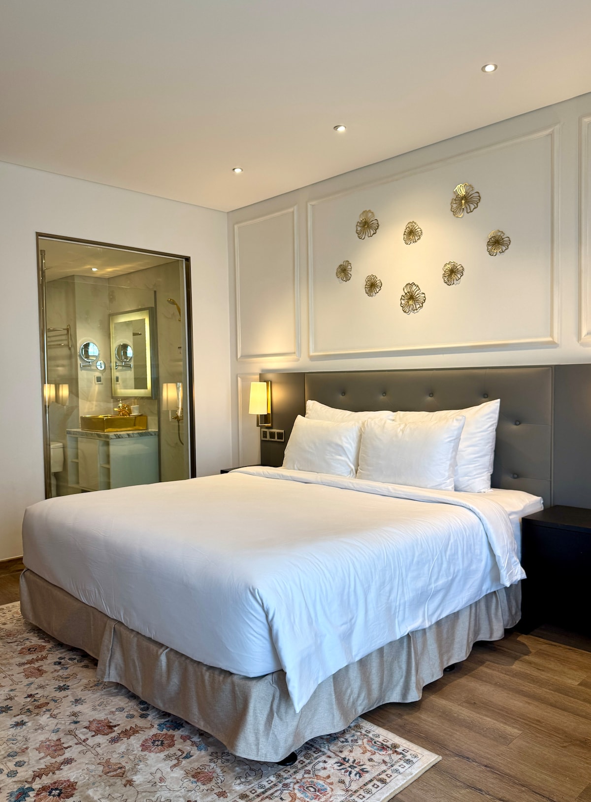 A well-appointed bedroom features a spacious bed with crisp white linens. Decorative wall elements add a touch of elegance. Soft lighting highlights the room’s neutral tones, while a glass-enclosed bathroom is visible in the background, providing a sense of modern luxury.