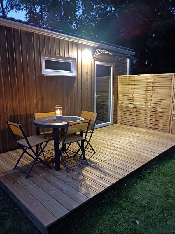 A wooden terrace features a round table with two folding chairs and a lantern, providing illumination. The exterior is lined with wooden panels, contributing to a rustic feel. A door leads into the interior space, framed by a light entering from a nearby source.