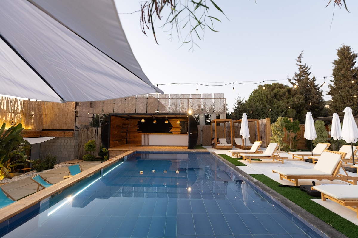 A spacious outdoor pool area features a glistening water surface under ambient lighting. Comfortable lounge chairs are arranged beside the pool, alongside a bar area that blends into the natural surroundings. Umbrellas provide shade while nearby greenery adds a tranquil touch to the setting.