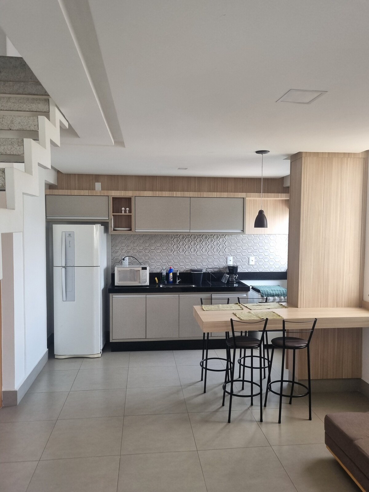 A modern kitchen area is equipped with a white refrigerator and a black countertop. Bar stools are positioned at a wooden dining table. The backsplash features a textured geometric pattern, and natural light enters through a nearby window, illuminating the space.
