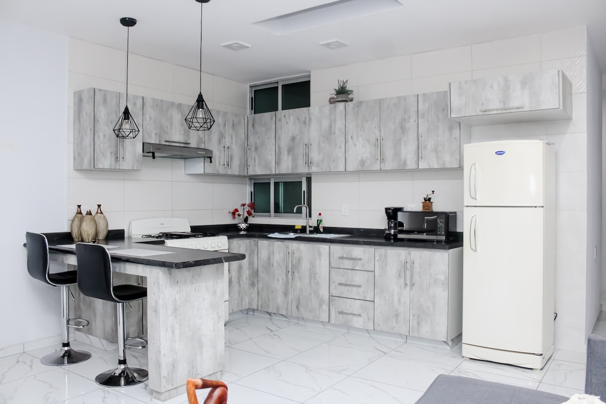 A modern kitchen features sleek gray cabinetry and a contrasting black countertop. Two bar stools sit at the counter, and essential appliances including a refrigerator, stove, and coffee maker are visible. Natural light illuminates the space through a window, enhancing the design.