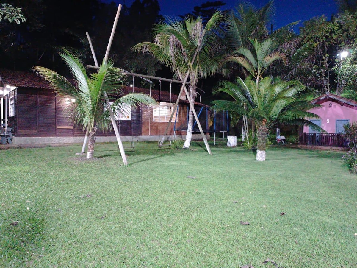 A spacious outdoor area is shown, featuring several palm trees and a manicured lawn. In the background, two wooden houses are visible, alongside a playground structure designed for children. Soft lighting accentuates the greenery and structures, contributing to a serene environment.