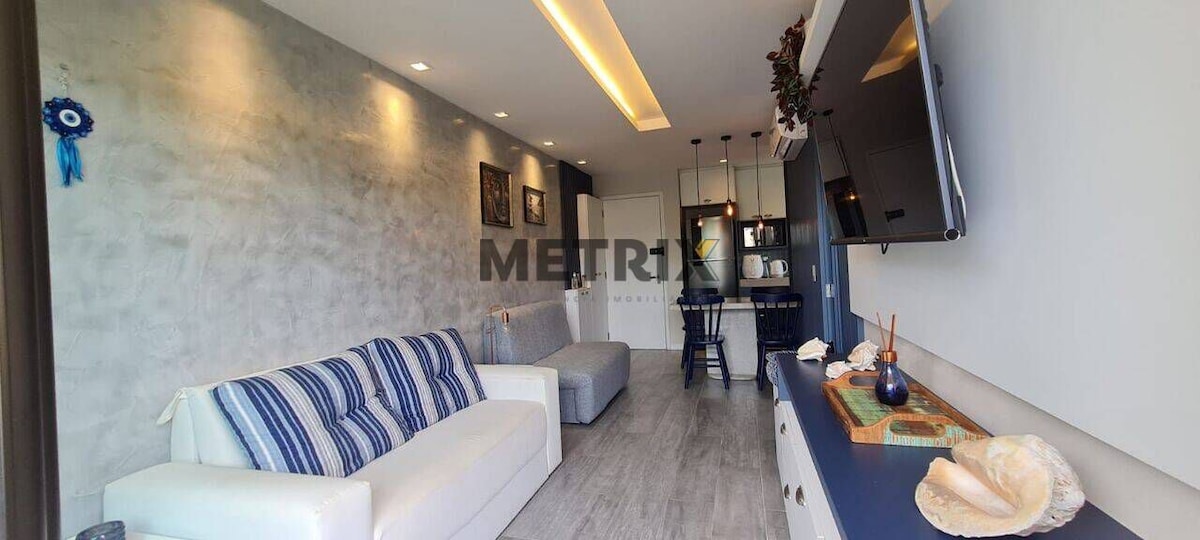 A modern living area features a light-colored sofa with blue and white striped cushions, complemented by a gray sofa bed. A wall-mounted TV is positioned above a decorative table that displays seashells. Soft lighting enhances the calming atmosphere, while a kitchen area is visible in the background.