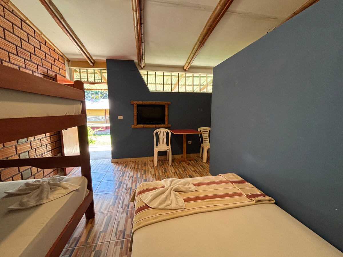 A simple and functional sleeping space features a bunk bed alongside a spacious bed. A small table is paired with two chairs, while a TV is mounted on the wall. Natural light enters through the window, casting a soft glow across the room.