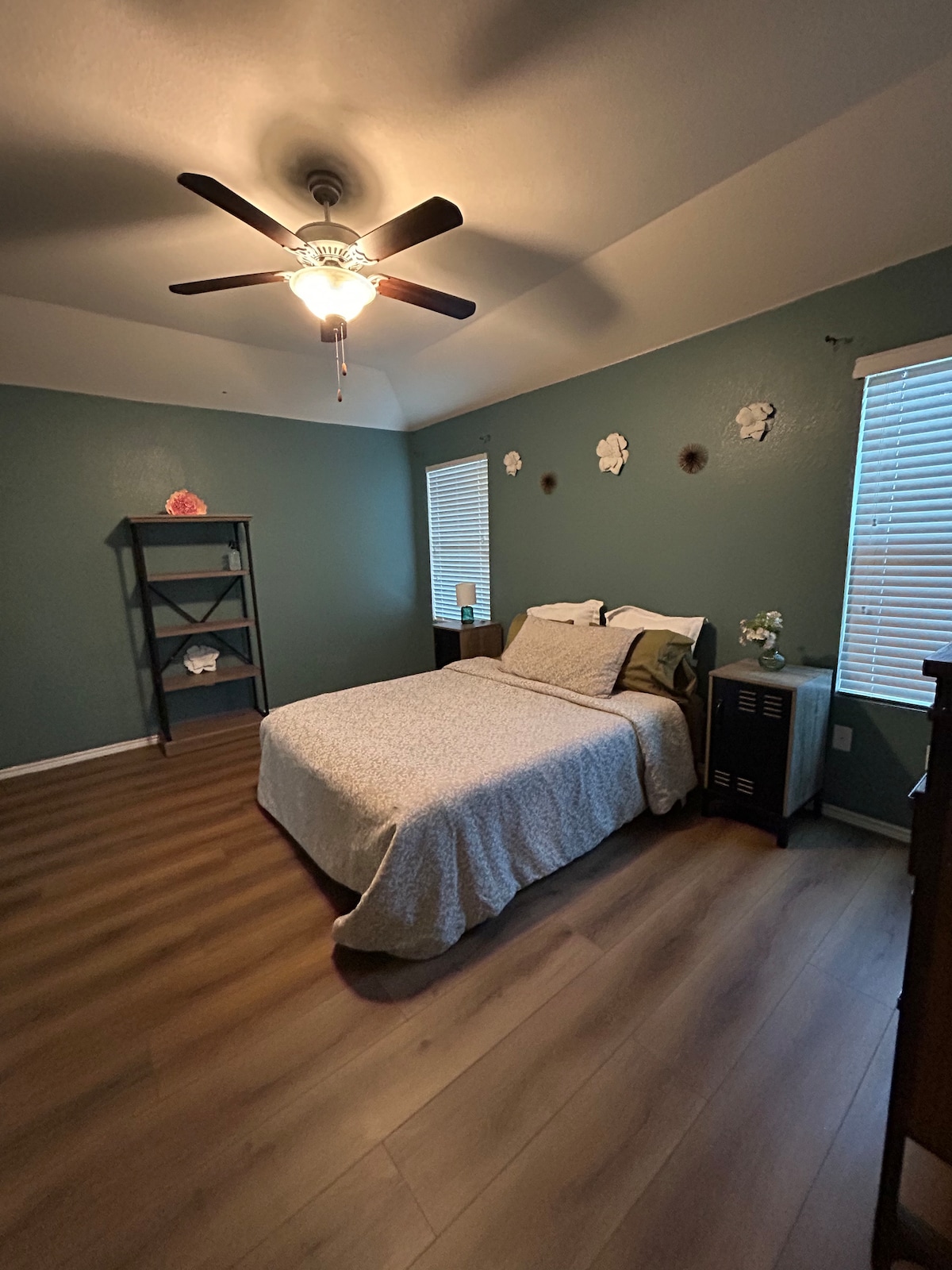 A calm bedroom features a spacious bed covered with a light-colored quilt. Two windows allow natural light to fill the space, accented by light green walls and floral wall decor. A wooden shelf stands in one corner, complementing the warm wooden flooring.