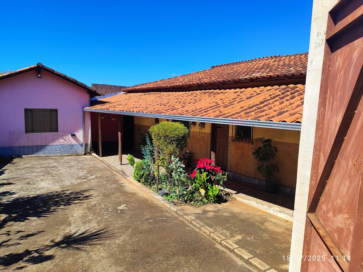 A single-story building with a terracotta roof is positioned alongside a landscaped pathway filled with vibrant flowers. A neighboring pink structure and a clear blue sky are visible, adding a bright backdrop to the outdoor setting.