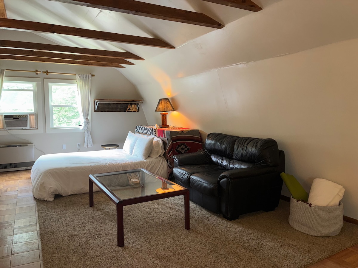 A spacious studio cottage is presented, featuring a comfortable bed and a cozy leather sofa. Natural light enters through two windows, complemented by decorative shelving and a lamp providing soft illumination. The carpeted floor adds warmth to the overall ambiance of the space.