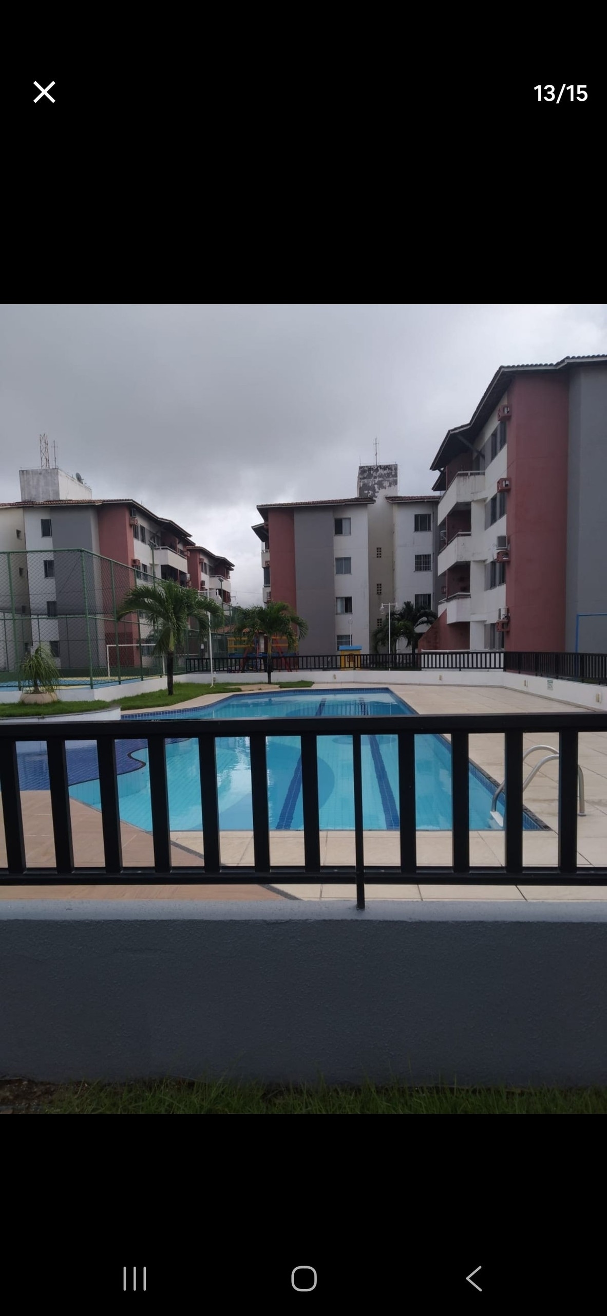 A clear view of a well-maintained swimming pool is presented, surrounded by landscaped areas and palm trees. The pool is framed by modern apartment buildings, showcasing a mix of light and dark exterior colors. The atmosphere is calm, with cloudy skies overhead.