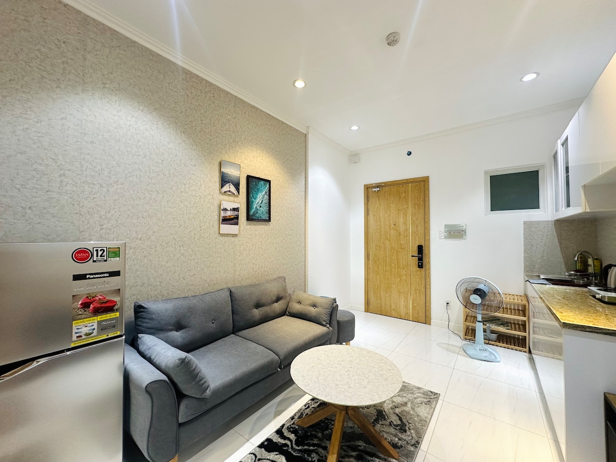 A modern living area features a gray sofa positioned across from a round table on a patterned rug. An upright fan is present, and a refrigerator is visible nearby. Wall decor includes three framed images, and natural light enters through a window on the far side.