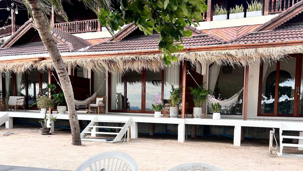 A row of charming Southern Thai-style bungalows features thatched roofs and large glass windows. Potted plants add greenery, while hammocks are visible on the porches. Sandy grounds and a few white chairs contribute to a relaxed outdoor setting with a clear view of the sea in the background.