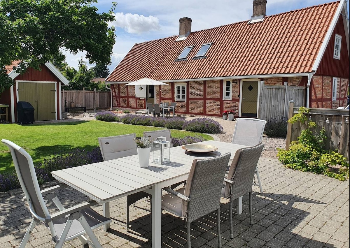 A well-furnished outdoor dining area is presented, featuring a large table with several chairs. The serene garden is surrounded by colorful flowers and green grass, complemented by a charming red structure in the background. A barbecue grill is visible to the side.