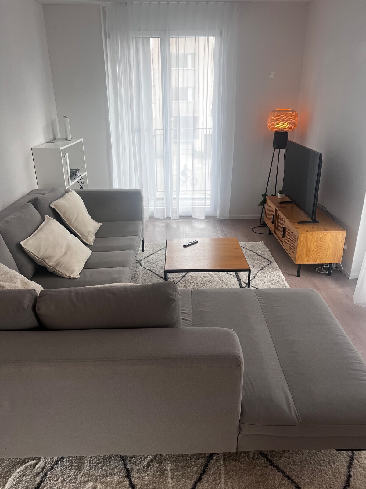 A light-filled living area is furnished with a wide, gray sectional sofa adorned with white cushions. A small wooden coffee table is positioned in front, and a television set rests on a wooden media console. Sheer curtains cover a balcony door, enhancing the relaxed ambiance.