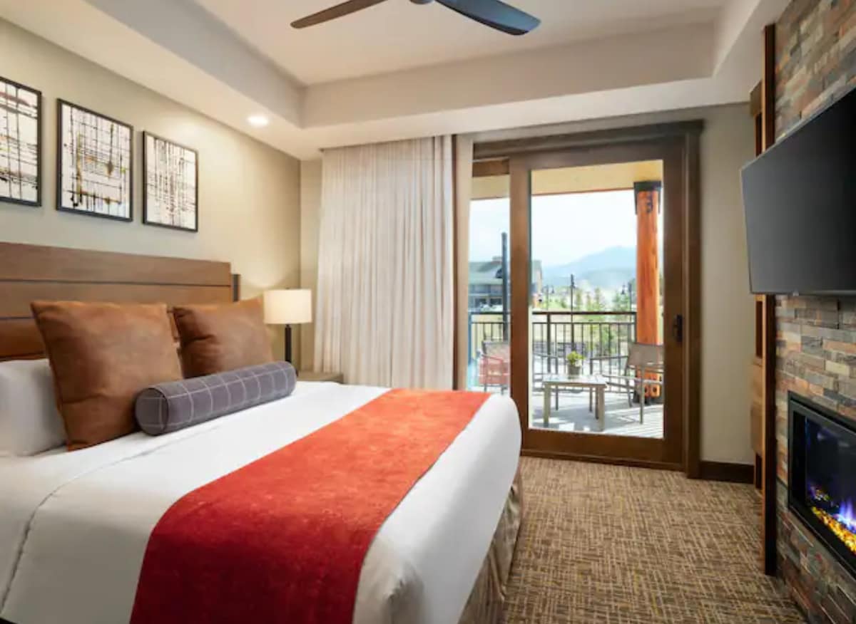 A cozy bedroom features a spacious bed dressed in white linens with orange and brown accent pillows. A modern fireplace is positioned below a wall-mounted TV. Natural light enters through large glass doors, which lead to a private balcony overlooking scenic mountain views.