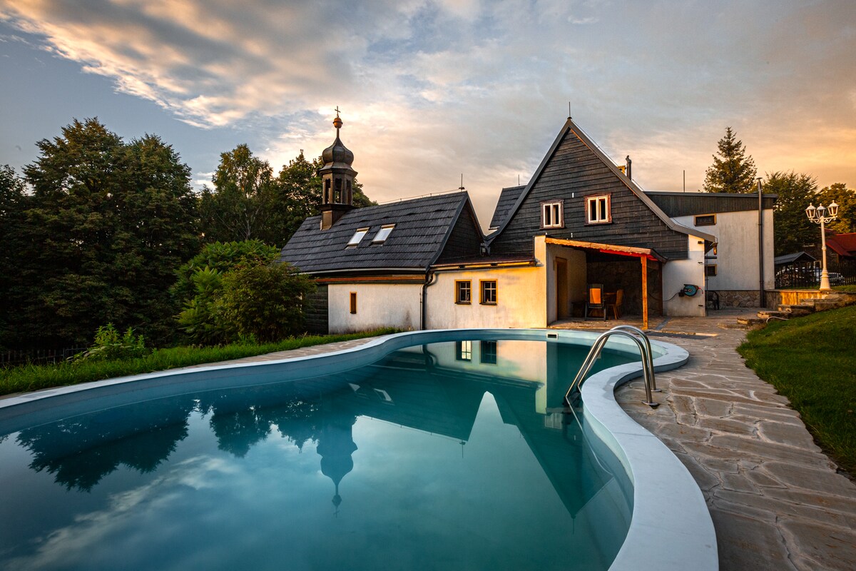 A swimming pool with a curved edge reflects the colors of the sunset, surrounded by lush greenery. Behind the pool, a traditional cottage with a sloped roof features small windows and an outdoor seating area, creating a serene atmosphere for relaxation.