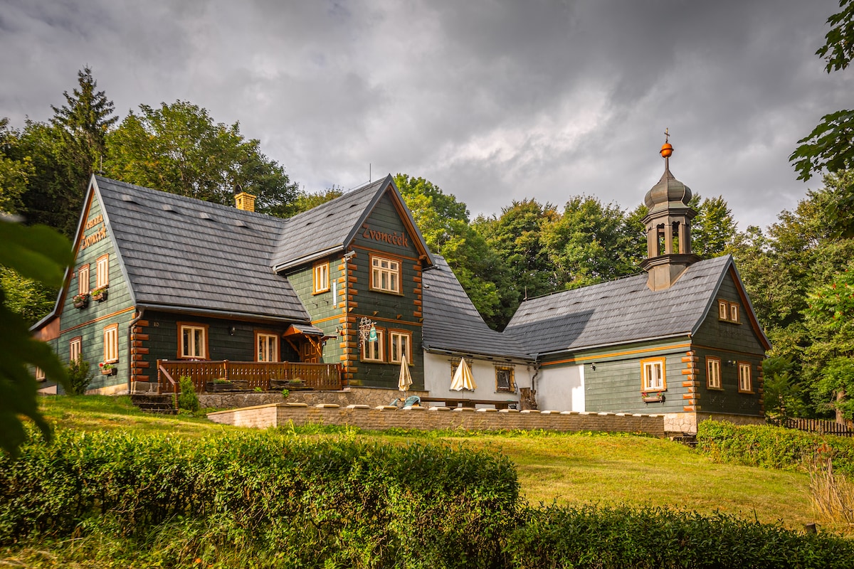An original cottage, constructed in 1781, is surrounded by lush trees and features a distinctive clock tower. The building's wooden façade displays green and brown tones, with large windows framed by light trim. A spacious terrace with umbrellas is visible, offering outdoor seating.