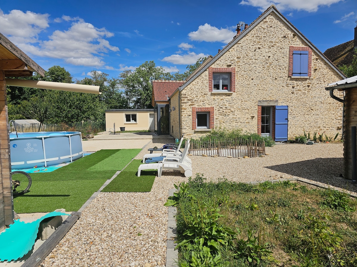 The exterior view showcases a charming two-story stone house with blue shutters. Sun loungers are arranged on green turf near a swimming pool. The surrounding yard features gravel pathways and patches of greenery, creating an inviting space for relaxation.
