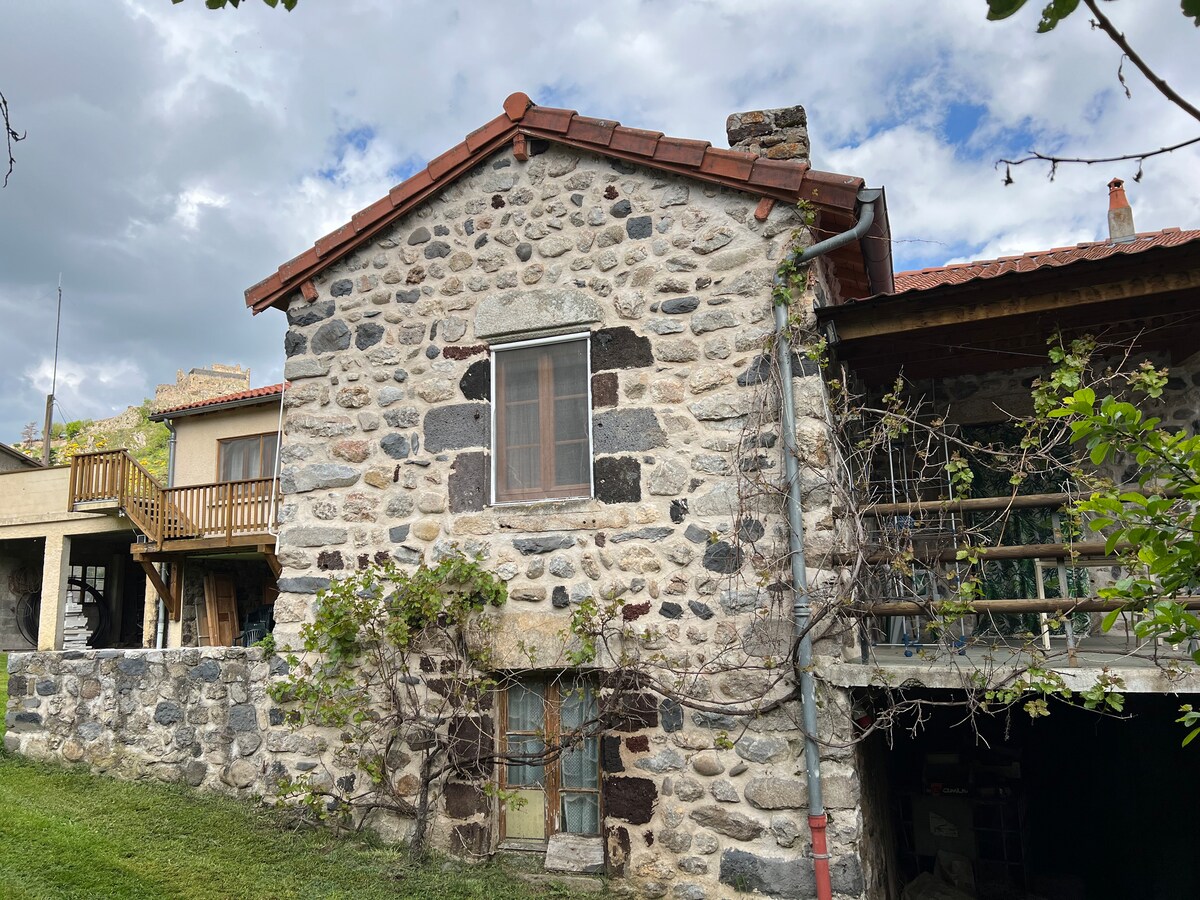 A rustic stone farmhouse is displayed, featuring a traditional red-tiled roof. The façade showcases various sizes of stones, adding texture. Green vines climb the side of the building, and wooden balconies are visible on the upper level, complementing the natural surroundings.