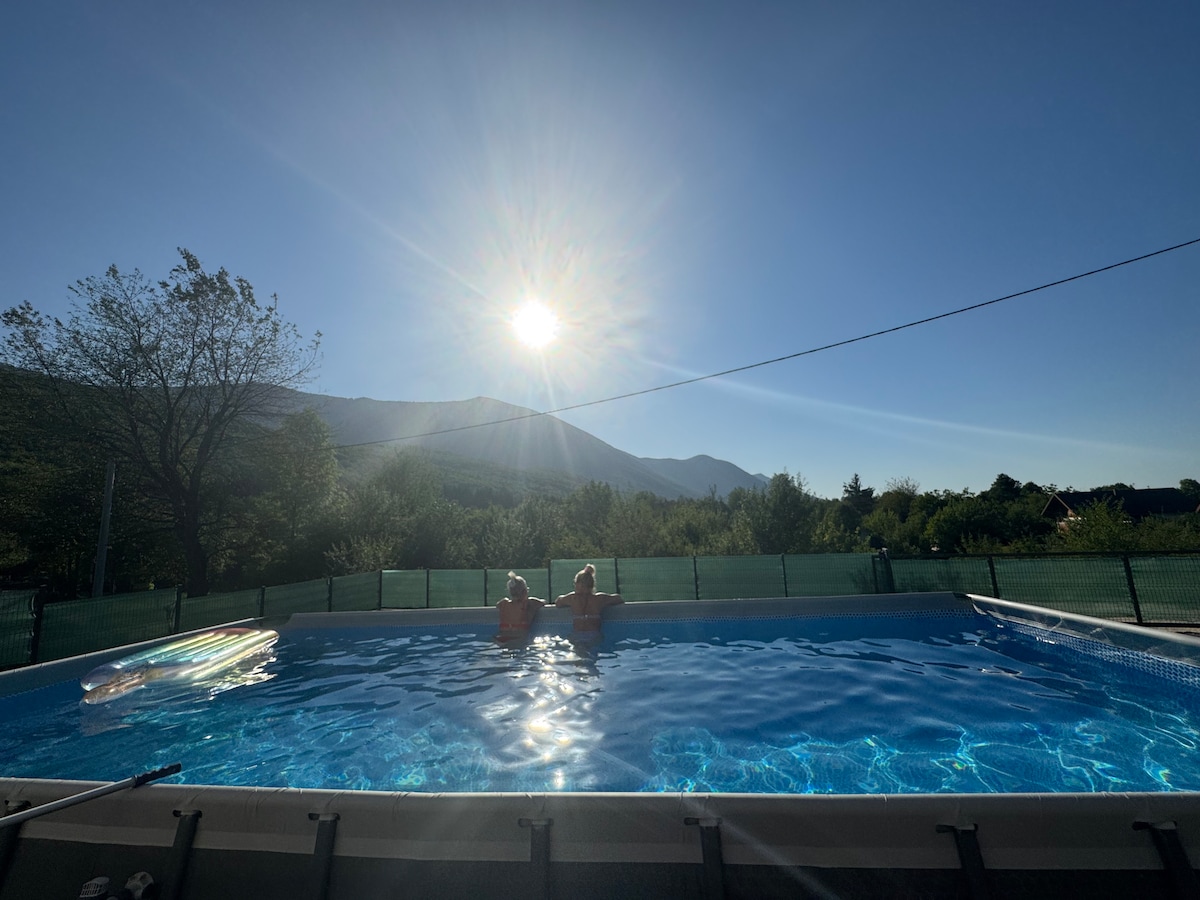 A swimming pool is visible, reflecting sunlight and surrounded by greenery. Two individuals are seated at the edge, facing distant mountains under a clear blue sky. A floating device rests in the water, adding to the serene atmosphere.