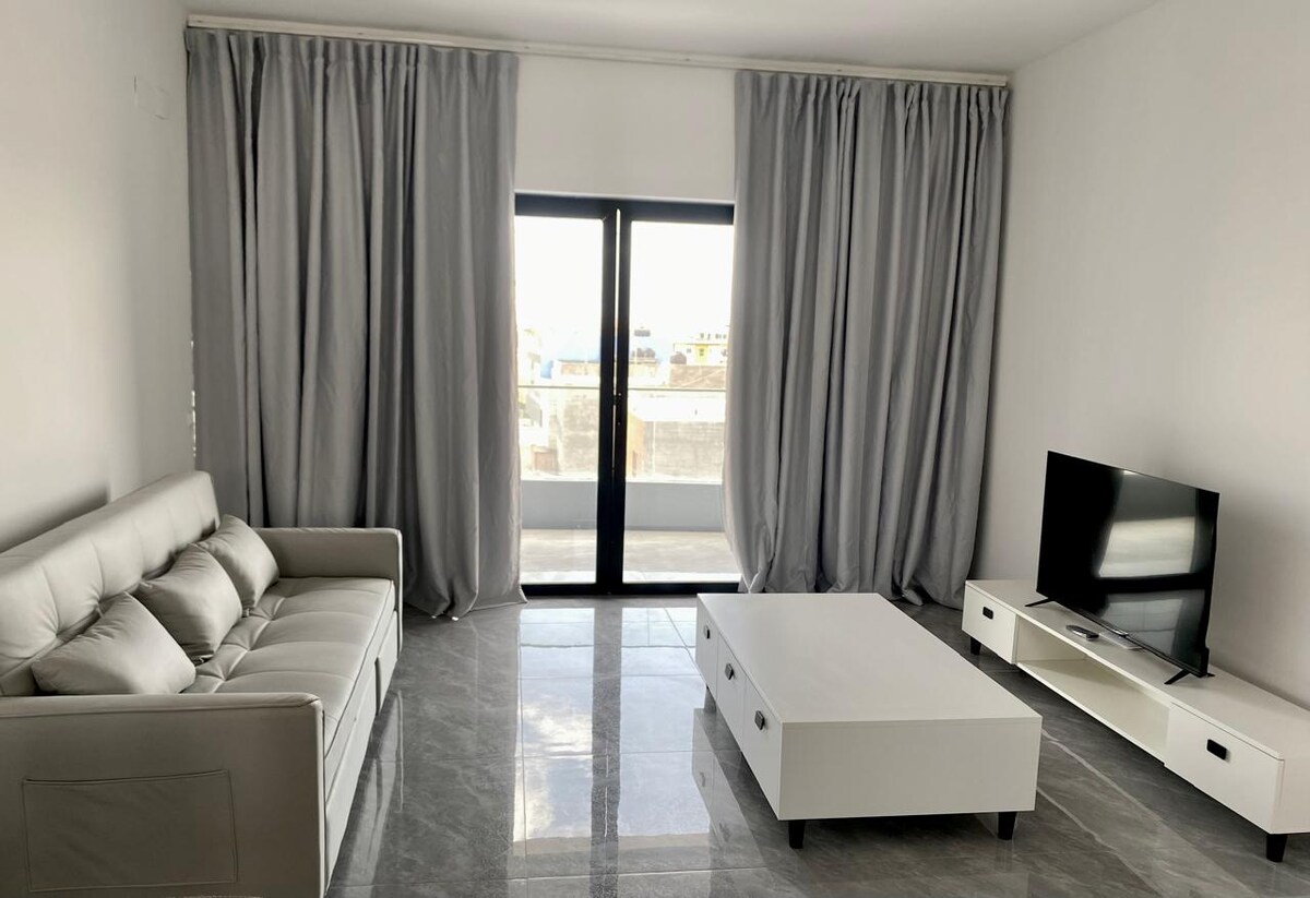 A living area is presented with a light-colored sofa positioned opposite a modern TV unit. Large glass doors provide natural light and lead to a balcony, while flowing gray curtains frame the windows. A low, white coffee table adds to the spacious feel.