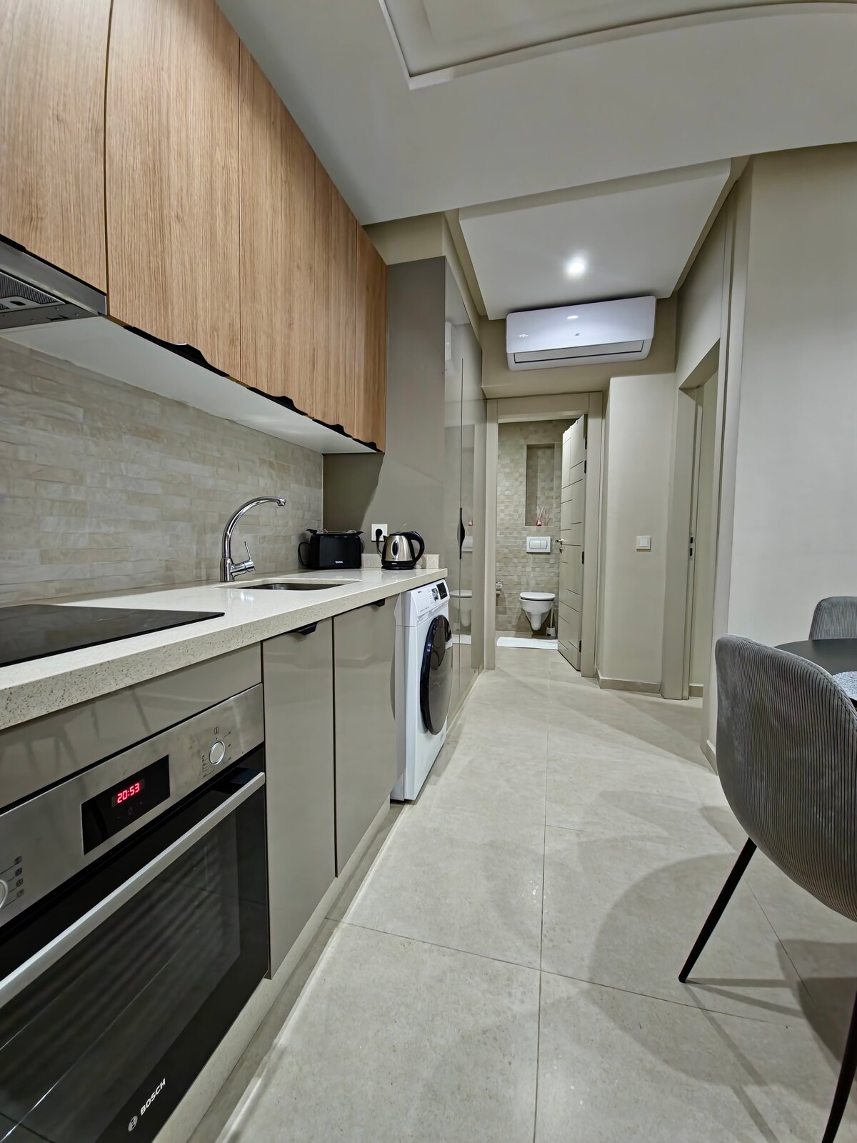 A modern kitchen features wooden cabinetry and sleek countertops. Essential appliances are visible, including an oven, stove, and washing machine. A small dining area is positioned nearby, with a round table and gray chairs. An open layout allows for easy movement within the space.