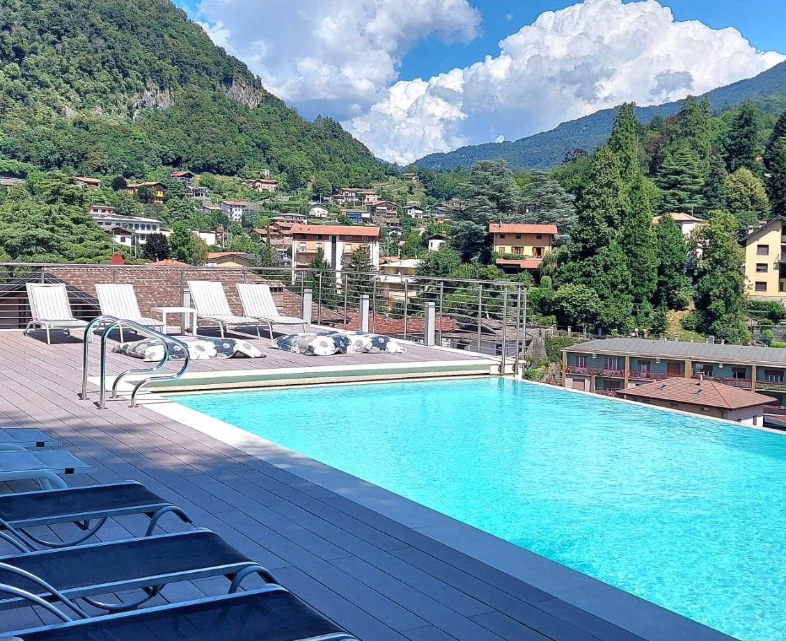 An inviting rooftop pool features a serene blue surface, framed by comfortable lounge chairs. The surrounding mountains and green trees create a calm backdrop, while the nearby buildings provide a glimpse of the local architecture under a partly cloudy sky.