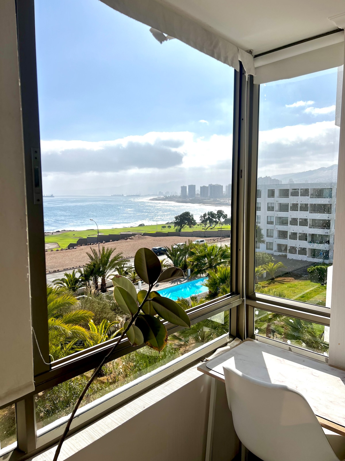 A wall of glass windows offers a view of the beach and distant city skyline. Lush greenery and a swimming pool are visible in the foreground, creating a bright and airy atmosphere within the space.