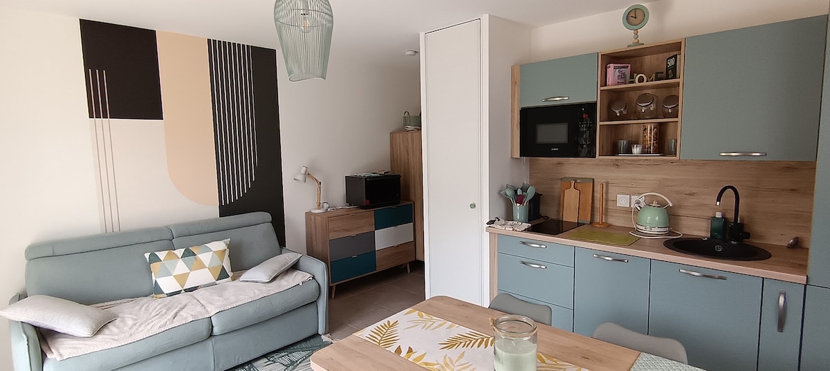 A cozy studio space features a light teal sofa adorned with patterned cushions. The kitchen area includes modern cabinetry, a microwave, and a sink. A wooden dining table is set with decorative elements, while a wall art piece adds a pop of color to the atmosphere.