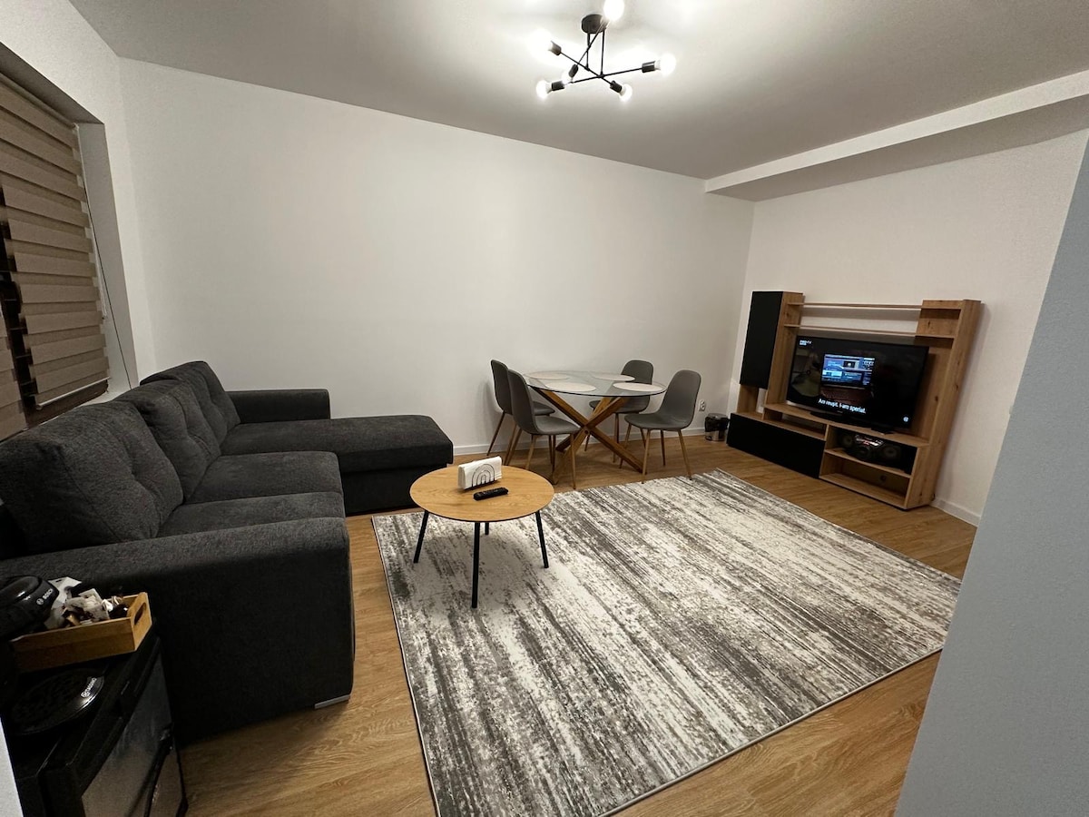 A modern living space showcases a comfortable grey sectional sofa and a round coffee table positioned on a textured area rug. A dining table with four chairs is visible nearby, alongside a media unit containing a television. Soft lighting is provided by a stylish overhead fixture.