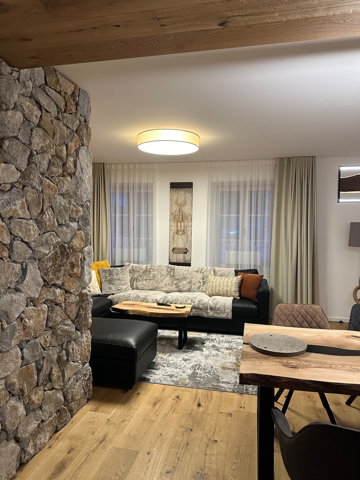 The living area showcases a sectional black sofa adorned with soft throws and cushions. A natural stone wall adds texture, while large windows provide ample light, dressed with elegant curtains. A wooden dining table is visible, accompanied by stylish seating.