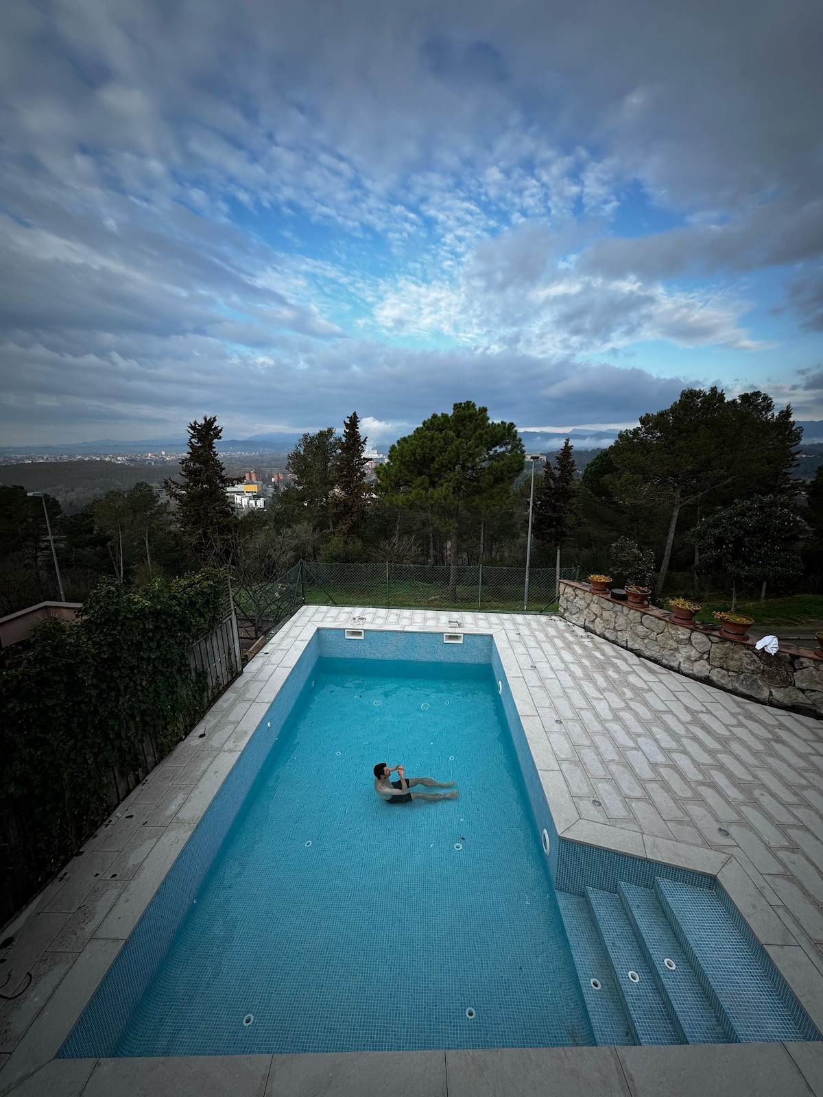 A private swimming pool is surrounded by stone walls and lush greenery, offering an expansive view of the landscape. A person floats serenely in the pool, reflecting the calmness of the environment. Bright blue skies with scattered clouds enhance the peaceful outdoor setting.