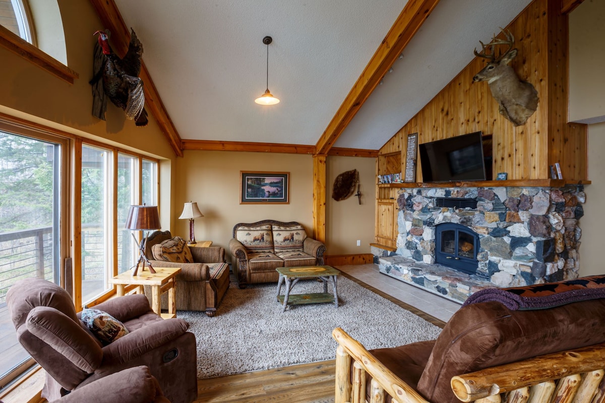 A spacious living room features a stone fireplace with a mounted deer head above it. A sofa and armchairs are arranged around a low coffee table on a light area rug. Large windows allow natural light to fill the room, with wooden beams accentuating the ceiling.