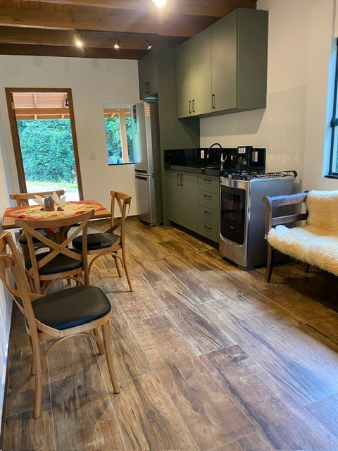 A kitchen area features modern cabinetry and appliances, including a refrigerator and stove. A dining table with four wooden chairs is positioned beside a cozy seating area. Natural light enters through a window, illuminating the wooden floor, complemented by a soft rug.