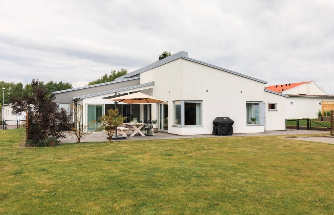 A modern single-story villa is set within a spacious garden. The outdoor seating area is shaded by an umbrella, with a dining table featuring several chairs. A barbecue grill is positioned nearby. Large windows provide views of the well-maintained grass and surrounding landscape.