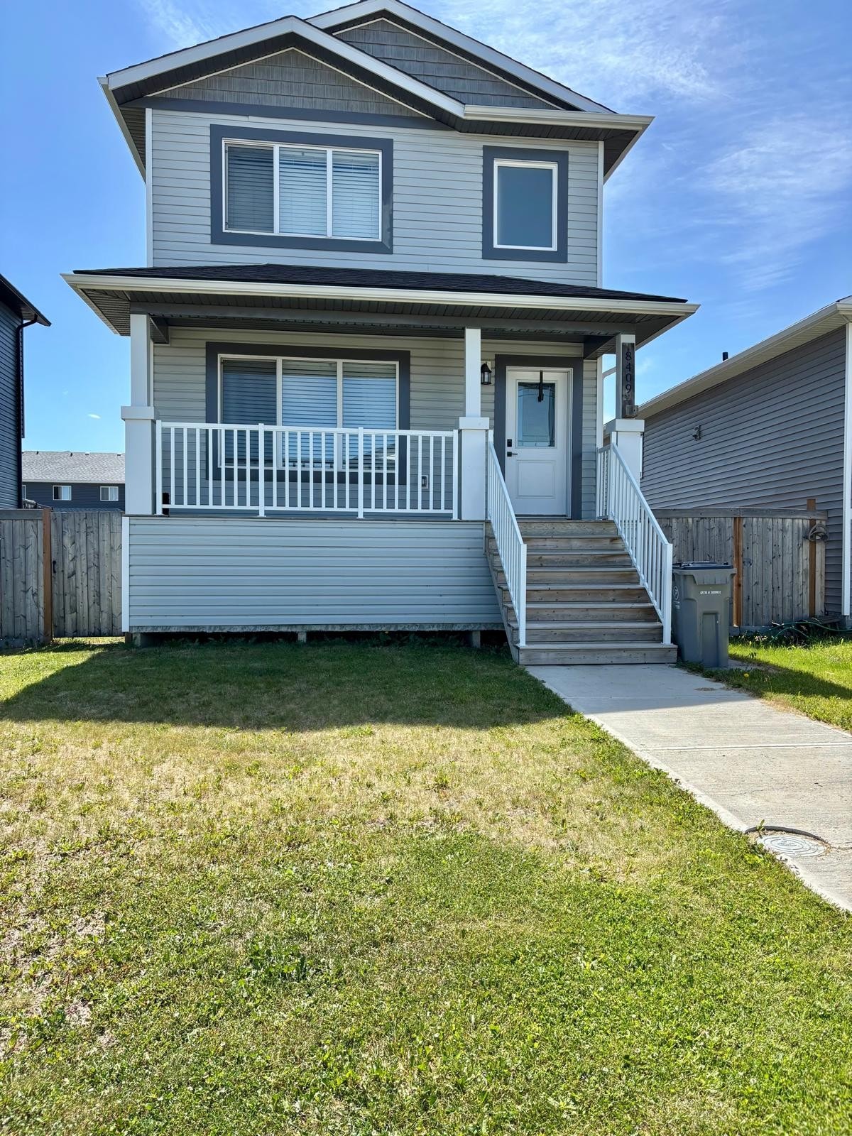 The exterior of a two-story house is shown, featuring a light grey façade and white trim. A welcoming front porch with white railings is accessed via a set of concrete steps. Lush green grass surrounds the home, with a fenced yard visible in the background.