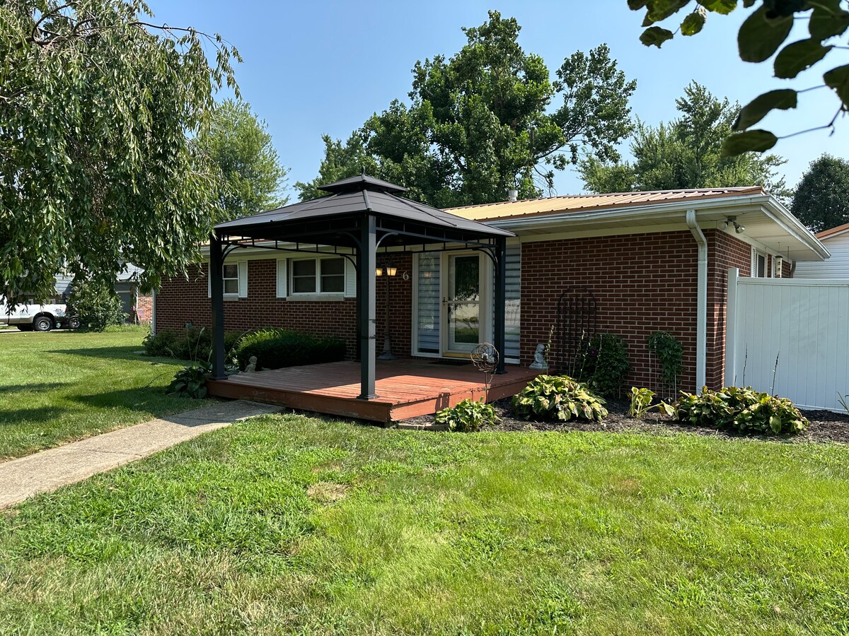 A welcoming front porch is featured, complete with a covered area supported by a sturdy structure. Lush greenery and blooming plants create a pleasant environment. The home is set against a backdrop of a well-maintained lawn, contributing to an inviting exterior.