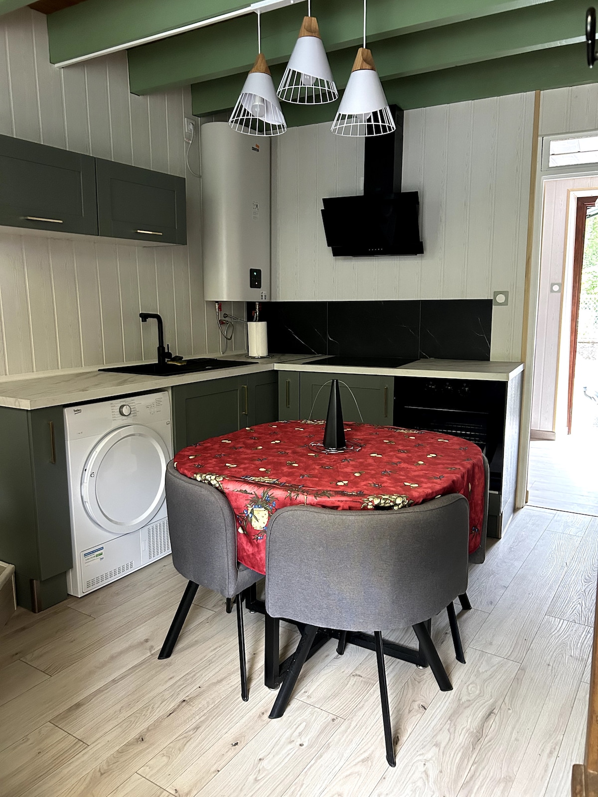 A compact kitchen area features modern cabinetry and appliances, including a washing machine and oven. A circular table with four chairs is set beneath pendant lighting, accented by a festive tablecloth. Natural light enters from a nearby doorway leading to an outdoor space.