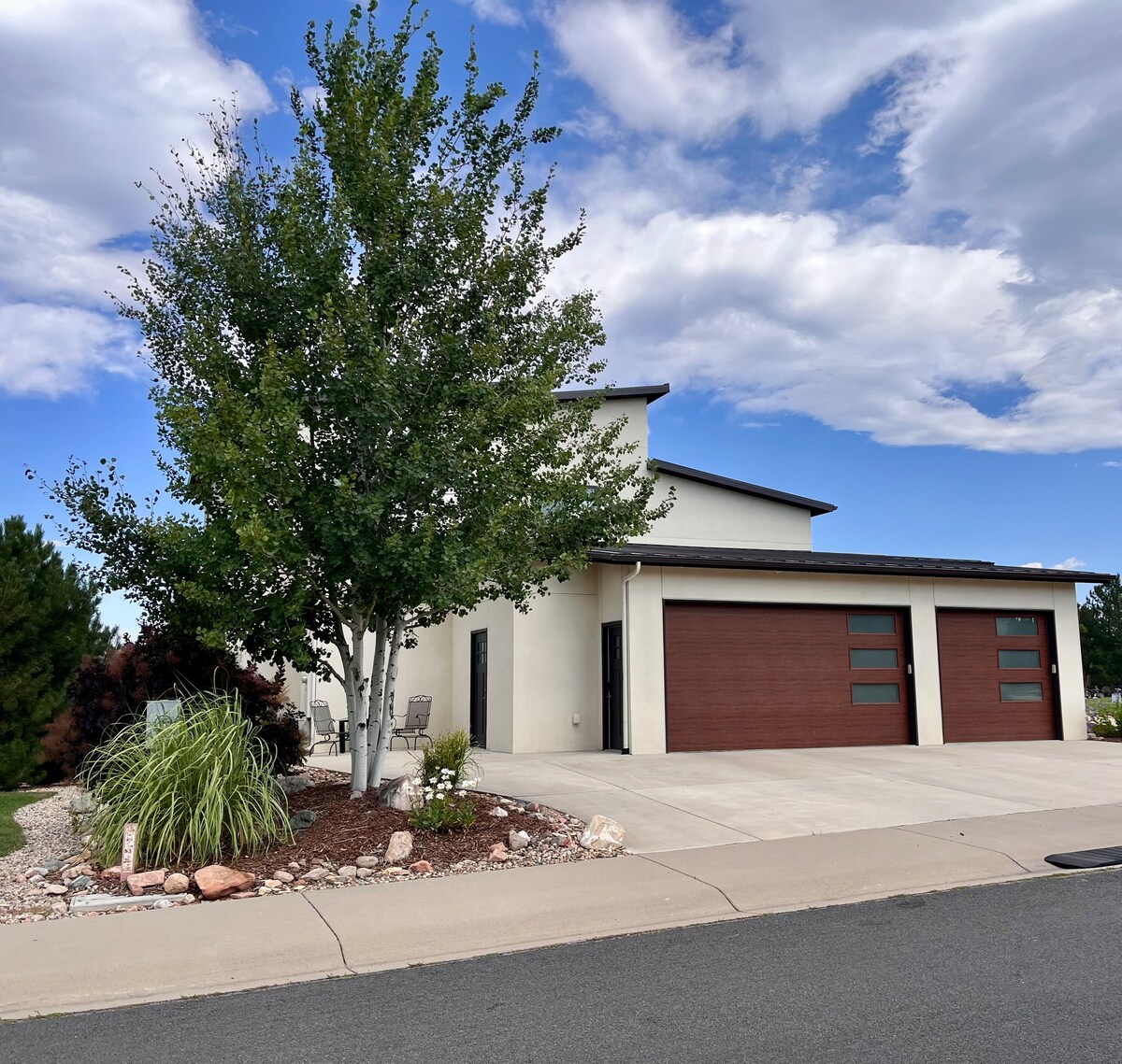 The exterior of a modern building is showcased, featuring a clean design and large garage doors. A tree with lush green foliage stands prominently in the front yard, alongside decorative landscaping elements. A small seating area is visible on the newly paved driveway.