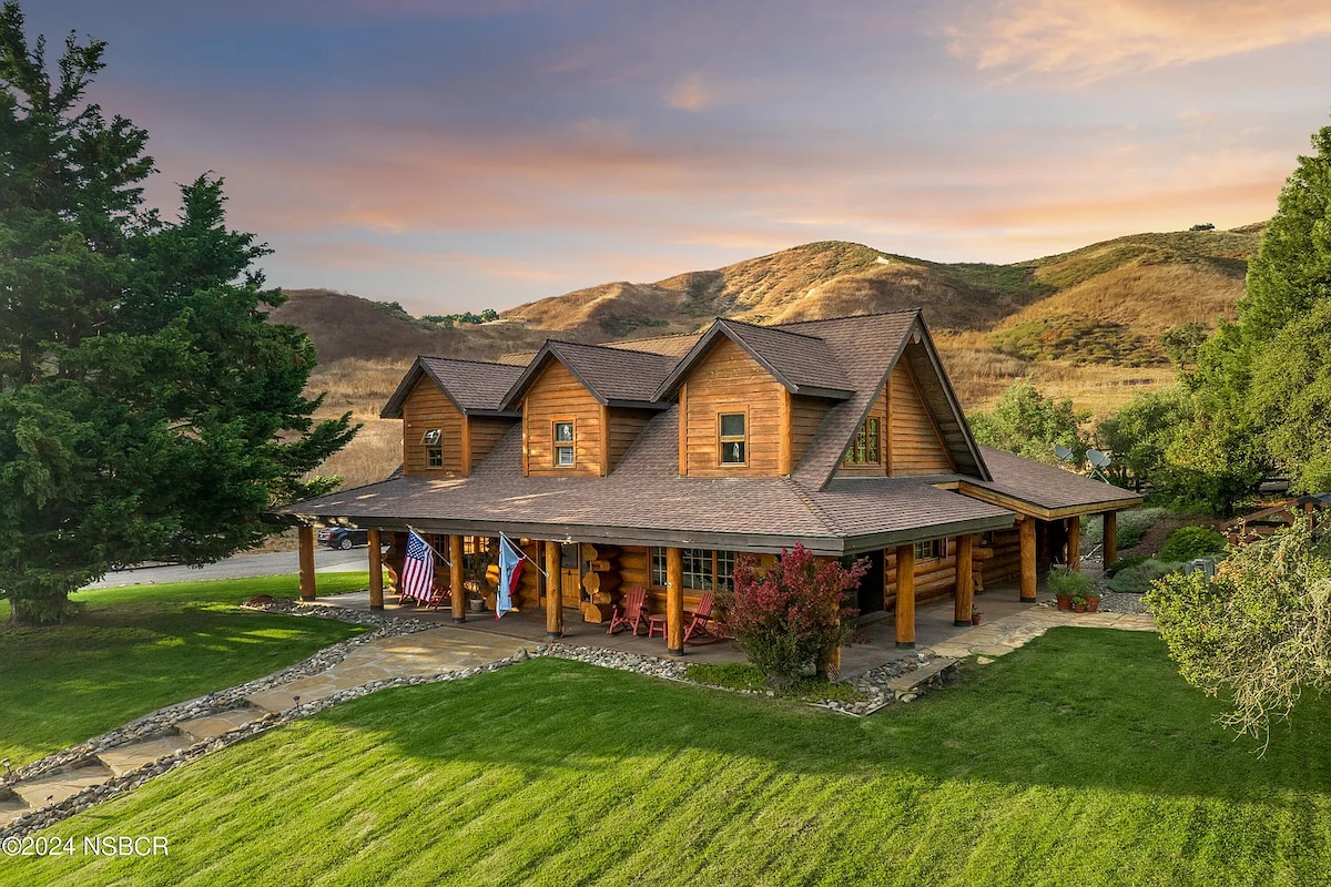 A spacious log cabin is framed by vibrant green grass and mature trees. The exterior features a covered porch with multiple entry points, and is designed with multiple gabled roofs. The backdrop reveals rolling hills under a colorful sunset sky, enhancing the natural setting.
