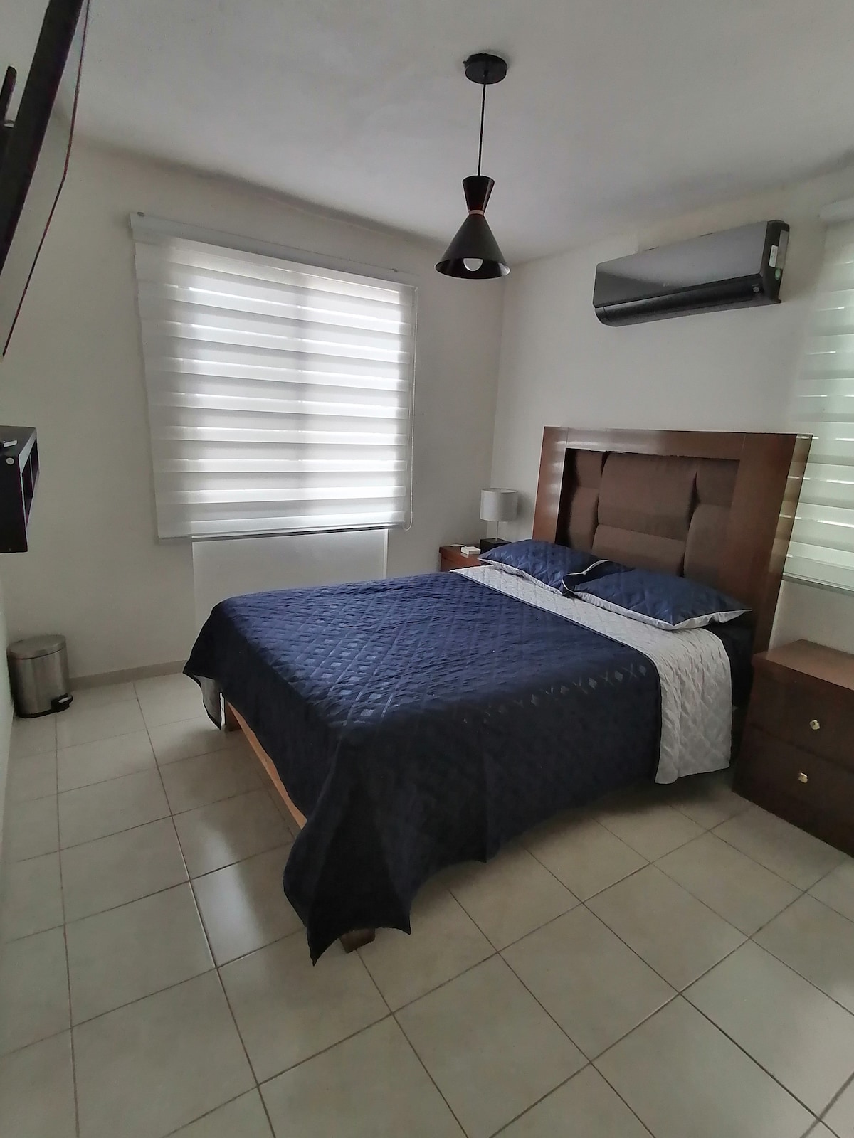 A well-appointed bedroom features a spacious bed with a navy blue coverlet. Natural light filters through sheer window treatments, complemented by blinds. A ceiling fan and air conditioning unit are visible, along with a nightstand and a television mounted on the wall.