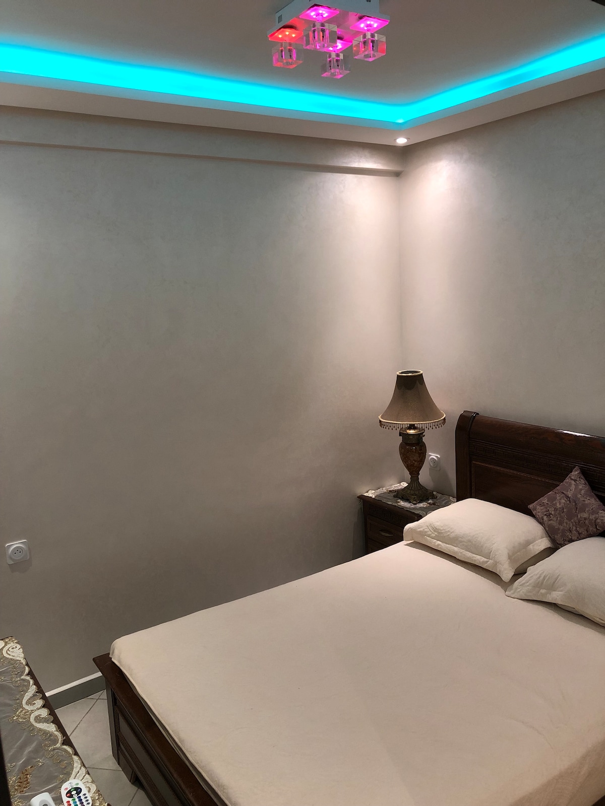 A cozy bedroom is presented with soft lighting from a ceiling feature that creates a subtle glow. A spacious bed with neutral bedding is positioned next to a nightstand with a decorative lamp. Light-colored walls complement the serene atmosphere of the room.