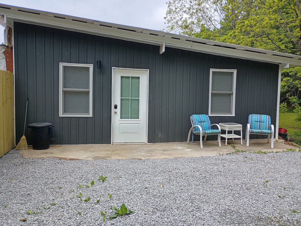 A gray exterior of a single-story building is showcased with two large windows and a central door featuring a glass panel. Two striped chairs and a small table are positioned on a concrete patio, surrounded by a gravel surface. Lush greenery is visible in the background.